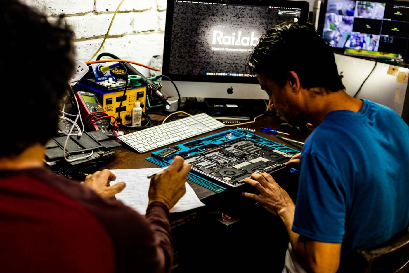 Educational setting: two people at a workbench reviewing a laptop motherboard with diagnostic tools and notes.
